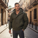 Man wearing a green jacket standing on a sunlit street with buildings in the background, Obroi