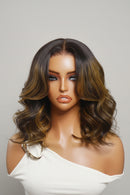 A wavy wig with dark brown roots and warm honey-brown highlights, styled on a mannequin head against a light gray background. The brand name 'obroi' is printed in the top left corner, Obroi