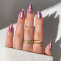 Hand with purple and clear nail polish wearing gold rings on a neutral background, Obroi