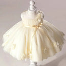 A cream-colored toddler party dress displayed on a white mannequin. The dress features a sleeveless bodice with a matching waist ribbon bow, a full tulle skirt, and decorative fabric flowers, Obroi