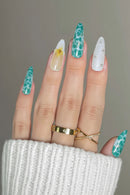 A close-up of a hand showcasing long, sculpted almond-shaped nails with various decorative designs: two teal and white marbled nails, one white nail with a yellow daisy, one white nail with silver rhinestones, and a purple nail at the corner. Gold rings are worn on the fingers, and a white knit sleeve is visible at the bottom, Obroi