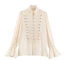 An off-white long-sleeve blouse with a high mock neck, pleated front panel, decorative cord details with gold-tone buttons, and flared bell sleeves, shown on a plain white background, Obroi