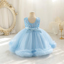 A light blue toddler girl's party dress displayed on a dress form mannequin in a bright, elegant room. The sleeveless dress features a scoop neckline with silver and blue sequin embroidery, a matching light blue satin bow at the waist, and a voluminous tulle skirt with ruffled trim, Obroi