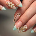Close-up of a hand with long almond-shaped nails decorated in a pastel French manicure style, featuring mint green tips, glossy pale pink bases, white floral motifs, gold metallic accents, and small pearl details, Obroi