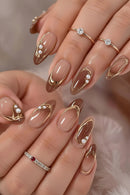 A close-up of hands showcasing elegant almond-shaped nail art with a glossy nude base, metallic gold curved line designs, small white pearl clusters, and delicate gold rings on the nails, Obroi