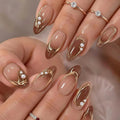 A close-up of hands showcasing elegant almond-shaped nail art with a glossy nude base, metallic gold curved line designs, small white pearl clusters, and delicate gold rings on the fingers, Obroi