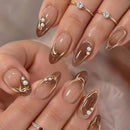 A close-up of hands showcasing elegant almond-shaped nail art with a glossy nude base, metallic gold curved line designs, small white pearl clusters, and delicate gold rings on the fingers, Obroi