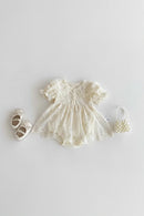 A baby girl's cream-colored lace dress with short flutter sleeves and a button-up front, styled in a flat lay with matching cream baby shoes and a small pearl handbag on a light gray background, Obroi
