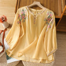 Yellow embroidered blouse on a wooden hanger with a wooden chair and flowers in the background, Obroi