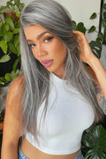 A woman wearing a long, straight gray wig with silver highlights, styled with a natural-looking lace part. She is wearing a white ribbed top and posing in front of a background of green houseplants, Obroi