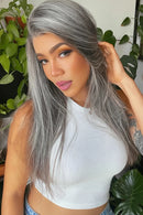 A woman wearing a long, straight gray wig with silver highlights, styled with a natural-looking lace part. She is wearing a white ribbed top and posing in front of a background of green houseplants, Obroi