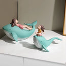 Two whimsical whale figurines on a white surface against a beige wall. One large mint-green whale has a girl figurine reclining on its back, and a smaller matching whale has a girl figurine sitting on its back, Obroi