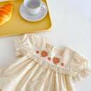 Beige dress with floral embroidery on a white surface, next to a yellow tray with a croissant and teacup, Obroi