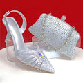 Silver high-heeled shoe with detailed embellishments and matching handbag on a red background, Obroi