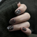 Hand with dark brown nail polish and black leopard print design on a dark background, Obroi