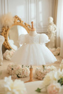 A white toddler girl's party dress with puff sleeves and a tulle skirt, displayed on a wooden dress form mannequin in a vintage-style room setting with a gold mirror and flowers, Obroi
