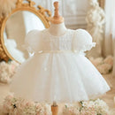 A white toddler girl's party dress with puff sleeves and a tulle skirt, displayed on a wooden dress form mannequin in a vintage-style room setting with a gold mirror and flowers, Obroi