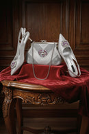 A coordinated women's formal accessories set displayed on a red velvet-covered wooden table against a dark wood paneled background. The set includes a silver rhinestone clutch handbag with a floral crystal brooch, a pair of pointed-toe slingback pumps in silver with matching crystal brooches, and a pair of pointed-toe pumps in silver with pink crystal embellishments, Obroi