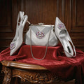 A coordinated women's formal accessories set displayed on a red velvet-covered wooden table against a dark wood panel background: a silver rhinestone clutch handbag with a chain strap, a pair of pointed-toe slingback pumps, and matching pointed-toe pumps with jeweled floral embellishments on the toes, Obroi