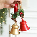 A hand holds two small Christmas bells hanging ornaments. One bell is metallic gold and the other is red, both with green pine branches, brown pinecones, red berries, and burlap hangers, Obroi