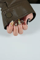 Close-up of hands with black nail polish and gold floral designs, wearing a brown leather jacket, Obroi