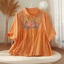 Orange blouse with floral embroidery on a white pedestal in a bright room, Obroi