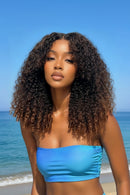 A woman wearing a Obroi curly hair extension with dark brown to black finger coil ends, set against a blue ocean background. An inset in the bottom right corner shows a close-up of the textured coil ends, Obroi