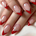 A close-up of a hand with long stiletto-shaped nails featuring a glossy nude-pink base, bright red tips, and a small silver rhinestone V-accent near the cuticle, Obroi
