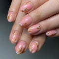 A close-up of manicured fingernails with a glossy light pink base, delicate white floral motifs, and metallic gold leaf accents, shown on a hand against a soft gray background, Obroi