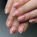 A close-up of manicured fingernails with a glossy light pink base, delicate white floral motifs, and metallic gold leaf accents, shown on a hand against a soft gray background, Obroi