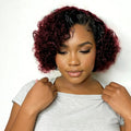 Woman with burgundy and black curly hair wearing a gray off-shoulder top on a white background, Obroi