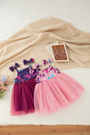 Two toddler girls' tutu dresses laid flat on a cream fabric surface with a book and small flowers as props. One dress is dark purple with butterfly print and magenta tulle skirt, with matching bow headbands, Obroi