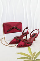 A pair of deep red satin pointed-toe slingback heels with jeweled embellishments, displayed with a matching red satin clutch bag on a white block and a gold chain strap, against a white draped background, Obroi