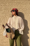 A woman wearing a cream pleated long-sleeve blouse with decorative front buttons, olive green pants, a red patterned headscarf, and sunglasses, holding a Gucci shoulder bag against a textured beige wall., Obroi