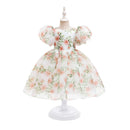 A white toddler girl's dress with a pink and green floral print, featuring puffy short sleeves and a full skirt, displayed on a white mannequin dress form against a white background, Obroi
