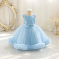 A light blue toddler girl's party dress displayed on a mannequin in a bright, elegant room. The dress features a lace bodice with floral embroidery, a matching blue bow at the waist, and a voluminous tulle skirt with a ruffled hem, Obroi