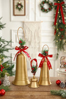 Three gold-toned Christmas bells of different sizes with jute hanging loops, red ribbon bows, pinecones, and greenery, displayed on a tabletop with holiday decorations in a softly lit indoor setting, Obroi