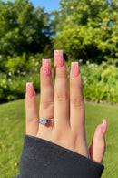 Hand with pink nail polish and a diamond ring against a green outdoor background, Obroi