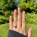 Hand with pink nail polish and a diamond ring against a green outdoor background, Obroi