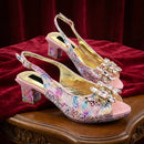 A pair of women's slingback high-heeled sandals with a pink and gold reptile-like pattern, embellished with rhinestones on the toe area and block heels, displayed on a red velvet cloth over a wooden table, Obroi