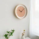 A minimalist round wall clock with a light wood frame and a dusty pink face, mounted on a white wall next to a small white vase with dried flowers and a green plant, Obroi