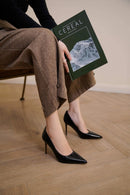 A person seated on a chair wearing brown textured trousers and black pointed-toe high heels, holding a dark green magazine with the word Obroi on the cover in an indoor room with a wood floor.