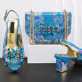 A matching women's fashion set by Queen Yan featuring a blue iridescent reptile-pattern handbag with gold chain straps and a blue rhinestone-embellished slingback sandal, shown on a white pedestal against a white curtain background, Obroi