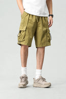 A person wearing olive green cargo shorts with side drawstrings, a white t-shirt, white socks, and beige hiking shoes, shown from the waist down against a light gray studio background, Obroi
