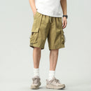 A person wearing olive green cargo shorts with side drawstrings, a white t-shirt, white socks, and beige hiking shoes, shown from the waist down against a light gray studio background, Obroi
