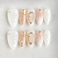 A set of decorative press-on nails arranged in two rows on a light background. The nails feature white, blush pink, and gold designs, including textured wing patterns, gold leaf accents, and pearl details, Obroi