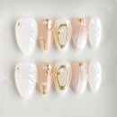 A set of decorative press-on nails arranged in two rows on a light background. The nails feature white, blush pink, and gold designs, including textured wing patterns, gold leaf accents, and pearl details, Obroi