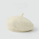 A textured cream-colored bouclé beret hat with a short top loop, photographed on a seamless light gray background, Obroi