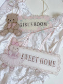 A styled flat-lay of baby girl nursery decor featuring a white lace baby romper, a teddy bear ornament, and two decorative signs reading 'GIRL'S ROOM' and 'SWEET HOME', Obroi