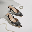 Pair of black embellished shoes on a light gray background with a pearl necklace, Obroi